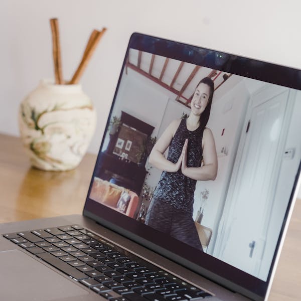 A woman participates in an online yoga class, seen on a laptop screen at home.